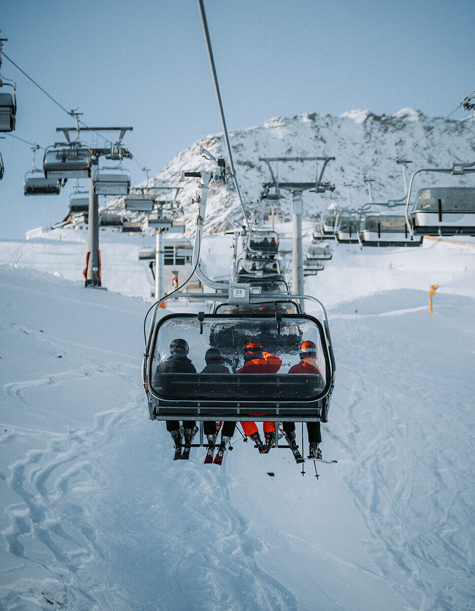 Vier Personen sitzen in einem Sessellift, der einen verschneiten Berg hinauffährt; unter ihnen sind Skispuren zu sehen, und im Hintergrund sind unter einem klaren Himmel weitere Sessellifte zu erkennen.