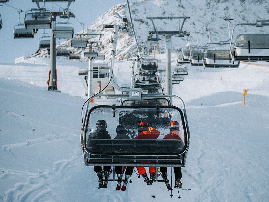 Vier Personen sitzen in einem Sessellift, der einen verschneiten Berg hinauffährt; unter ihnen sind Skispuren zu sehen, und im Hintergrund sind unter einem klaren Himmel weitere Sessellifte zu erkennen.