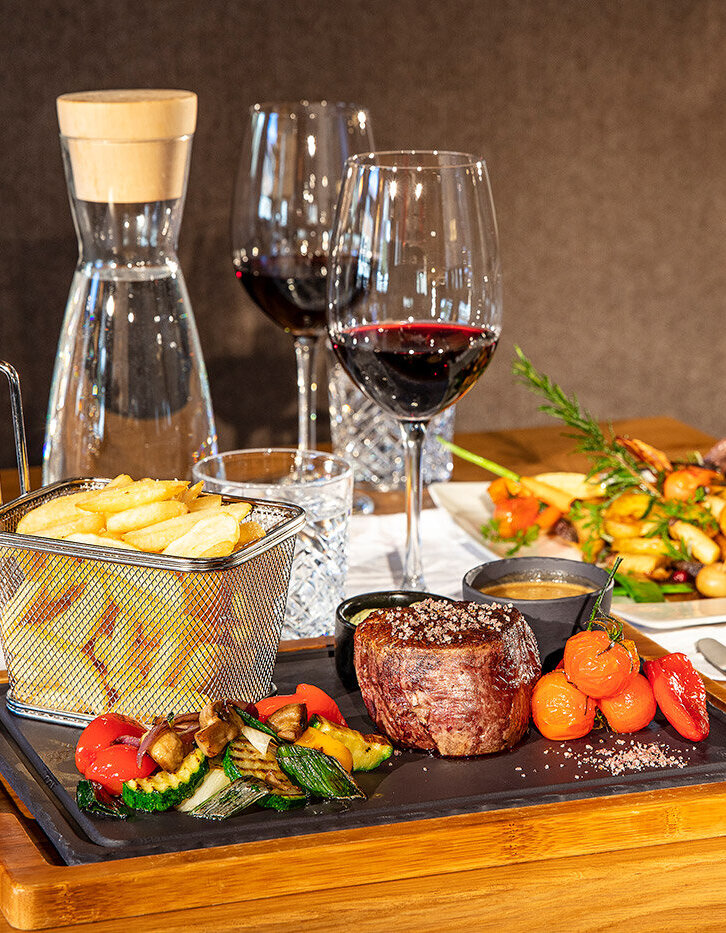 Auf einer Holzplatte liegen Steak, gegrilltes Gemüse, Kirschtomaten, Pommes frites in einem Korb und Soße. Zwei Gläser Rotwein, eine Wasserkaraffe und eine Kerze stehen auf einem gedeckten Esstisch, im Hintergrund ist ein weiteres angerichtetes Gericht zu sehen.