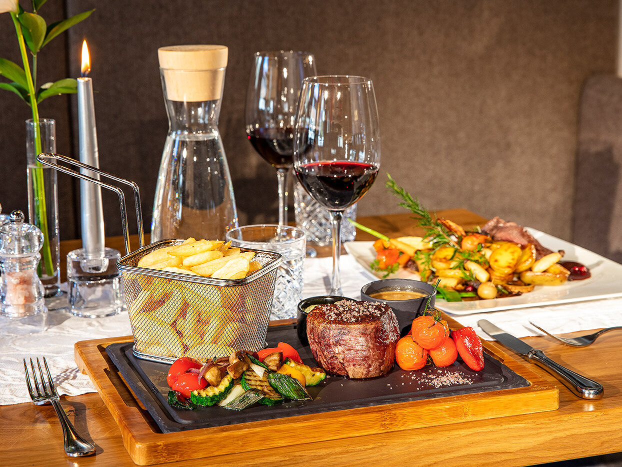 Auf einer Holzplatte liegen Steak, gegrilltes Gemüse, Kirschtomaten, Pommes frites in einem Korb und Soße. Zwei Gläser Rotwein, eine Wasserkaraffe und eine Kerze stehen auf einem gedeckten Esstisch, im Hintergrund ist ein weiteres angerichtetes Gericht zu sehen.