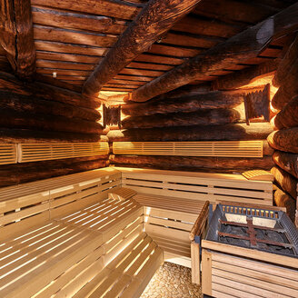Innenansicht einer rustikalen Holzsauna mit Blockwänden, gestuften Holzbänken, warmer Beleuchtung und einem mit Steinen gefüllten Ofen in einer Ecke. Der Boden ist mit Fliesen in Steinoptik ausgelegt.