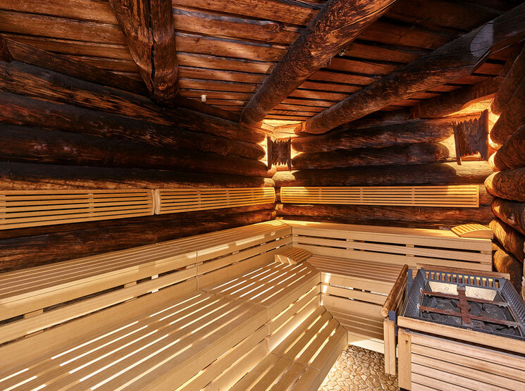 Innenansicht einer rustikalen Holzsauna mit Blockwänden, gestuften Holzbänken, warmer Beleuchtung und einem mit Steinen gefüllten Ofen in einer Ecke. Der Boden ist mit Fliesen in Steinoptik ausgelegt.