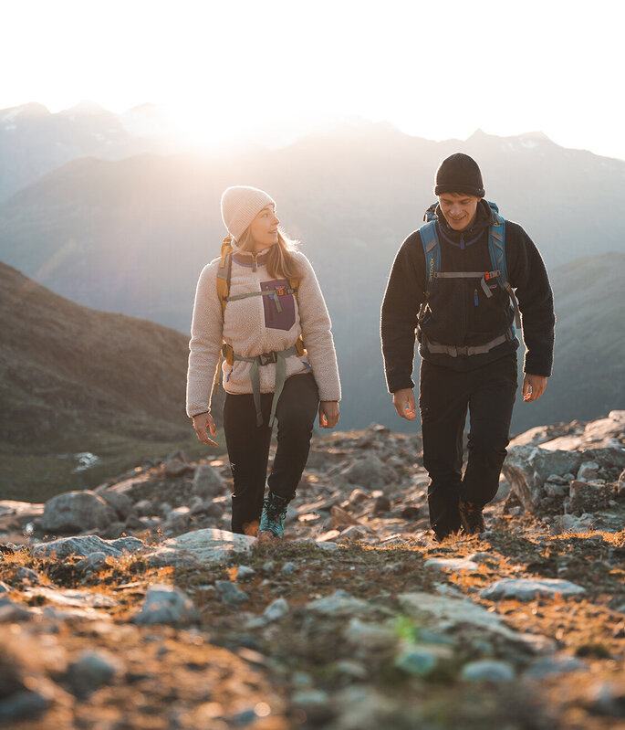 Zwei Menschen mit Rucksäcken und warmer Kleidung wandern bei Sonnenaufgang oder Sonnenuntergang auf einem felsigen Bergpfad, während im Hintergrund das Sonnenlicht über die fernen Gipfel strahlt.