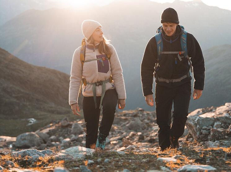 Zwei Menschen mit Rucksäcken und warmer Kleidung wandern bei Sonnenaufgang oder Sonnenuntergang auf einem felsigen Bergpfad, während im Hintergrund das Sonnenlicht über die fernen Gipfel strahlt.