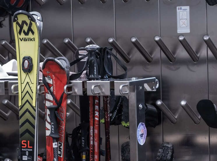Eine Nahaufnahme eines Skiraums, in dem Skier, Stöcke, Schuhe und sonstige Ausrüstung auf Metallregalen an einer Wand mit schräg angeordneten Haken verstaut sind.
