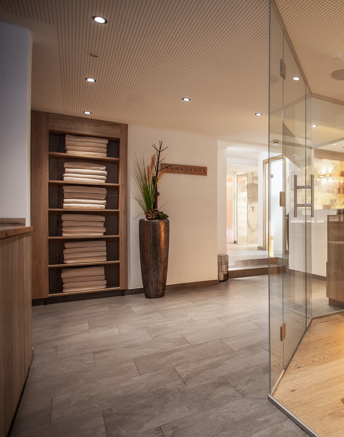 Ein modernes Spa-Interieur mit gefalteten Handtüchern auf Regalen, einer hohen Pflanze in einer Vase, Glaswänden und warmer Beleuchtung, die eine entspannte Atmosphäre schaffen.