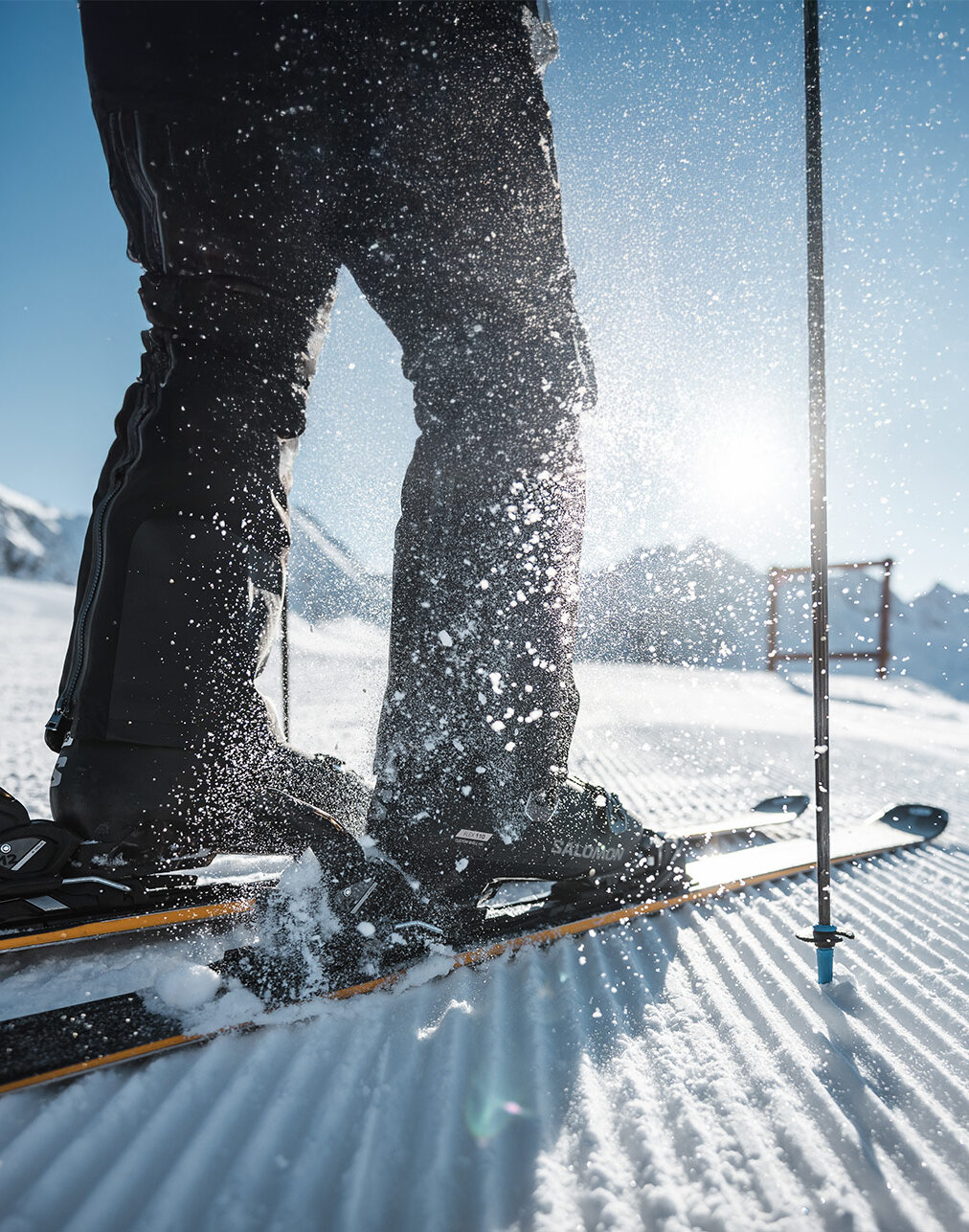 Nahaufnahme einer Person, die mit Skistöcken auf einem verschneiten Berghang steht, während Sonnenlicht durch den Schnee scheint und dieser hochspritzt; im Hintergrund sind Berge zu sehen.