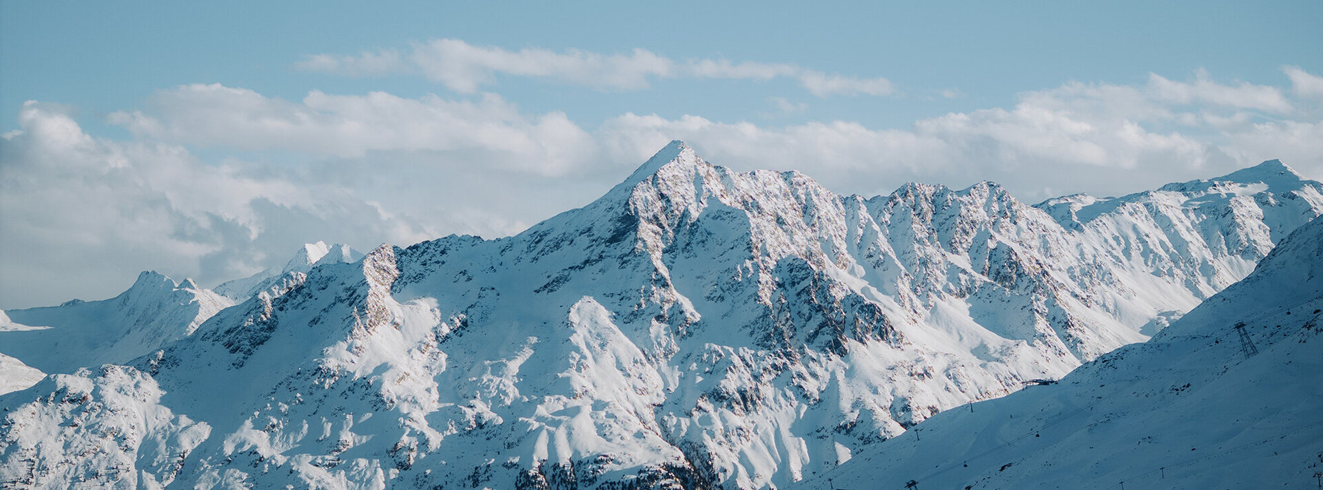 Eine schneebedeckte Bergkette unter blauem Himmel mit vereinzelten Wolken, die schroffe Gipfel und eine friedliche Winterlandschaft zeigt.