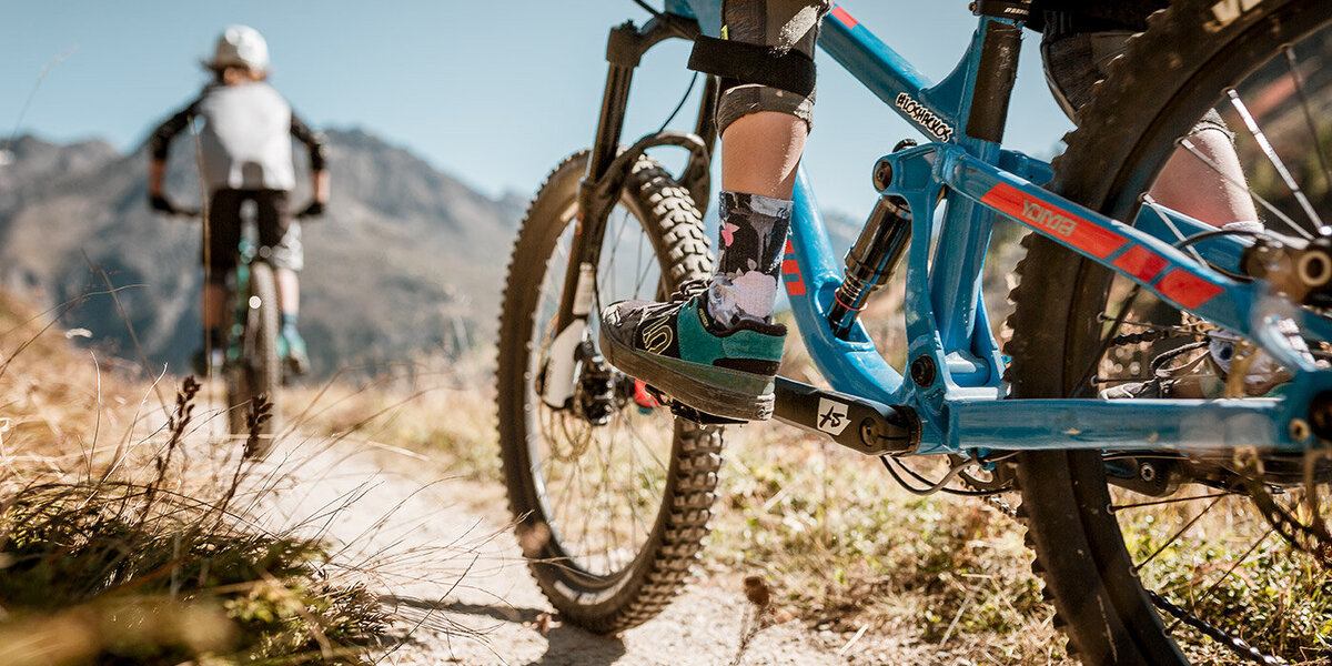 Zwei Personen fahren mit Mountainbikes auf einem Feldweg durch grasbewachsenes Gelände, im Hintergrund sind Berge zu sehen. Der Fokus des Fotos liegt auf den Beinen und den Fahrrädern, wodurch Bewegung und das Abenteuer in der freien Natur eingefangen werden.