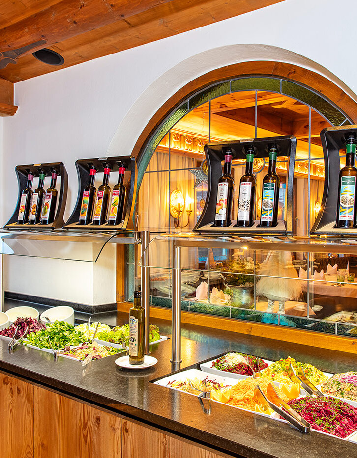 Eine Salatbar mit verschiedenen frischen Gemüsesorten und Blattgemüse in Schalen, darüber stehen Flaschen mit Öl und Essig, und durch ein Fenster sieht man draußen einen Parkplatz und Bäume.