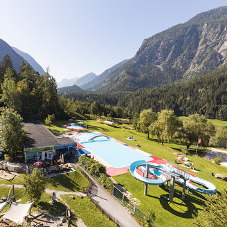Ein Freibad mit Rutschen und Liegeflächen, umgeben von Bäumen und Bergen unter strahlend blauem Himmel. Man sieht einige Menschen, die die Anlage genießen. Neben dem Poolbereich stehen Autos.