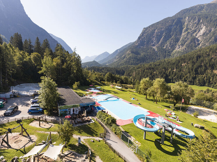 Ein Freibad mit Rutschen und Liegeflächen, umgeben von Bäumen und Bergen unter strahlend blauem Himmel. Man sieht einige Menschen, die die Anlage genießen. Neben dem Poolbereich stehen Autos.