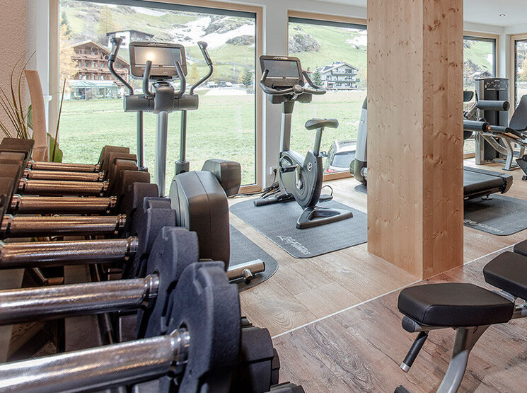 Ein modernes Fitnessstudio mit Holzelementen, ausgestattet mit einem Laufband, einer Hantelbank, einem Krafttrainingsgerät, freien Gewichten und großen Fenstern, durch die Tageslicht hereinströmt.