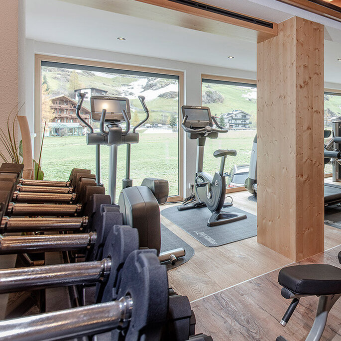Ein modernes Fitnessstudio mit Holzelementen, ausgestattet mit einem Laufband, einer Hantelbank, einem Krafttrainingsgerät, freien Gewichten und großen Fenstern, durch die Tageslicht hereinströmt.