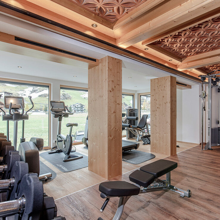 Ein modernes Fitnessstudio mit Holzelementen, ausgestattet mit einem Laufband, einer Hantelbank, einem Krafttrainingsgerät, freien Gewichten und großen Fenstern, durch die Tageslicht hereinströmt.