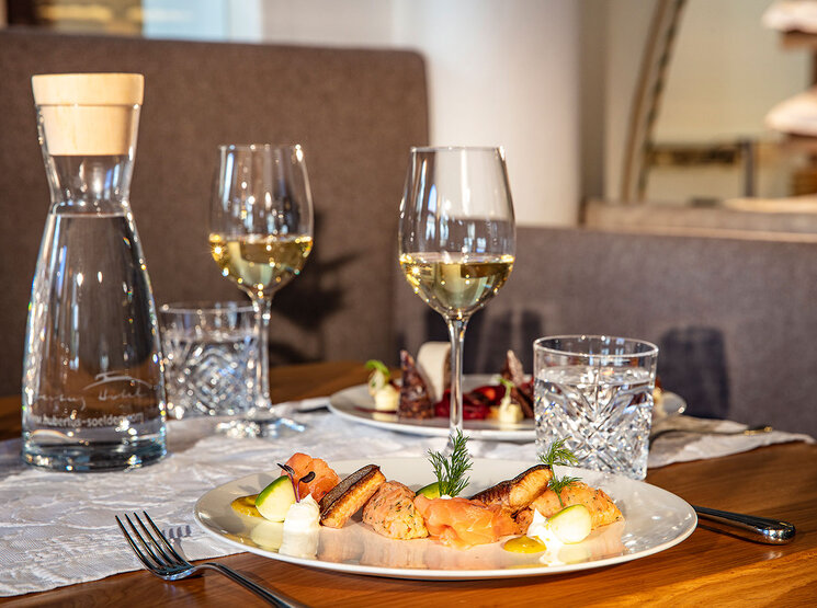 Ein für zwei Personen gedeckter Restauranttisch mit Tellern voller Gourmetgerichte, Gläsern mit Weißwein, Wassergläsern, einer durchsichtigen Karaffe und einem Tischläufer aus Leinen auf einem Holztisch. Im Hintergrund sind bequeme Sitzgelegenheiten und ein Teil der Restaurantinneneinrichtung zu sehen.