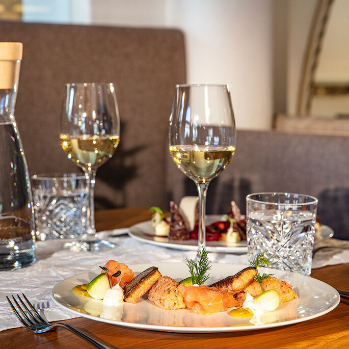 Ein für zwei Personen gedeckter Restauranttisch mit Tellern voller Gourmetgerichte, Gläsern mit Weißwein, Wassergläsern, einer durchsichtigen Karaffe und einem Tischläufer aus Leinen auf einem Holztisch. Im Hintergrund sind bequeme Sitzgelegenheiten und ein Teil der Restaurantinneneinrichtung zu sehen.