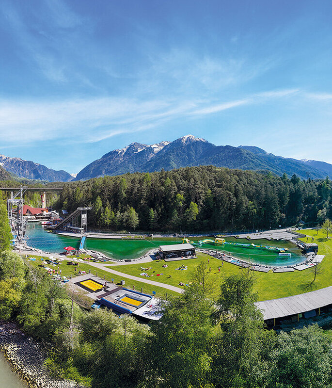Eine malerische Luftaufnahme der Area 47 mit grünen Rasenflächen, Schwimmbecken, Sonnenbadenden, Sportplätzen und bewaldeten Bergen im Hintergrund unter einem strahlend blauen Himmel.