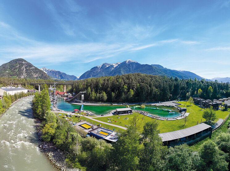 Eine malerische Luftaufnahme der Area 47 mit grünen Rasenflächen, Schwimmbecken, Sonnenbadenden, Sportplätzen und bewaldeten Bergen im Hintergrund unter einem strahlend blauen Himmel.
