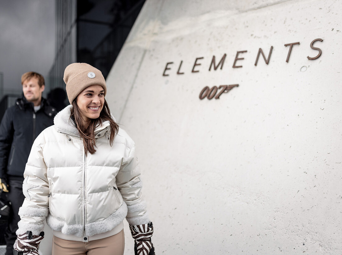 Eine lächelnde Frau in einer weißen Winterjacke und einer hellbraunen Mütze steht vor einem Gebäude, an dessen Wand „ELEMENTS 007“ steht. Im Hintergrund hält ein Mann in dunkler Kleidung Skiausrüstung in den Händen.