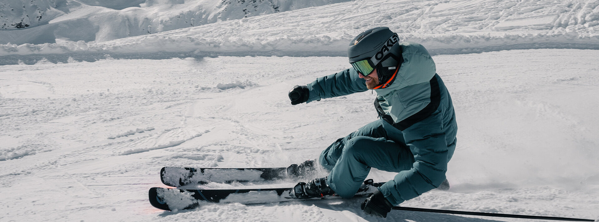 Ein Skifahrer in blauem Anzug und Helm macht eine scharfe Kurve auf einem verschneiten Berghang, wobei er eine Schneewolke aufwirbelt; im Hintergrund sind schneebedeckte Gipfel zu sehen.