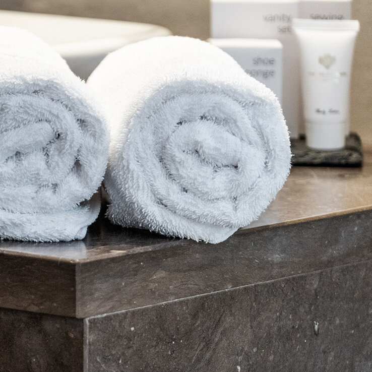 Zwei ordentlich aufgerollte weiße Handtücher liegen auf einer Marmorarbeitsplatte neben einem weißen Waschbecken, im Hintergrund sind verschwommene Toilettenartikel zu sehen, was auf ein sauberes, modernes Badezimmer hindeutet.