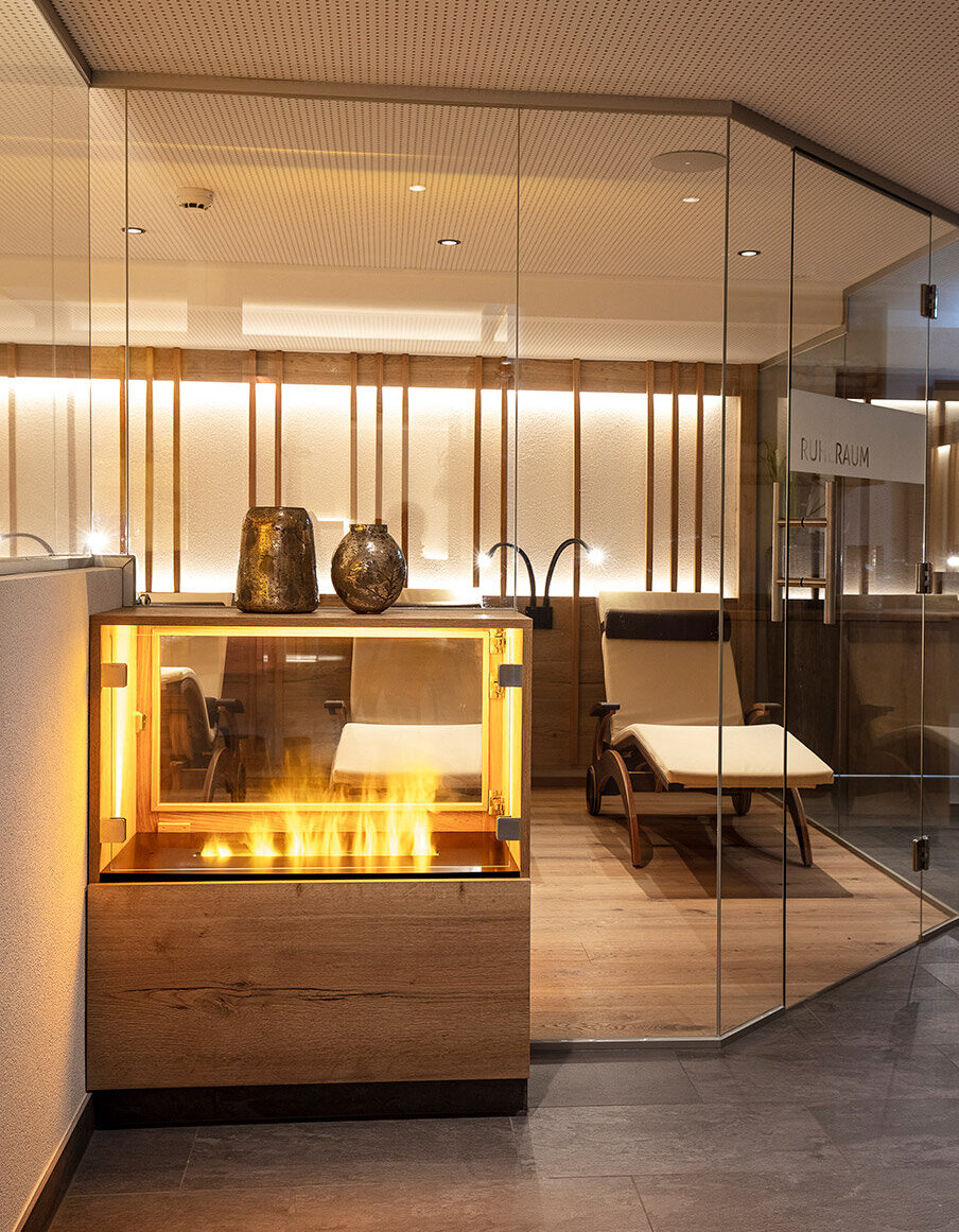 Moderner Wellness-Ruheraum mit Glaswänden, einem Liegestuhl aus Holz und einem dekorativen Kamin. Warme Beleuchtung und eine minimalistische Einrichtung schaffen eine ruhige, einladende Atmosphäre.