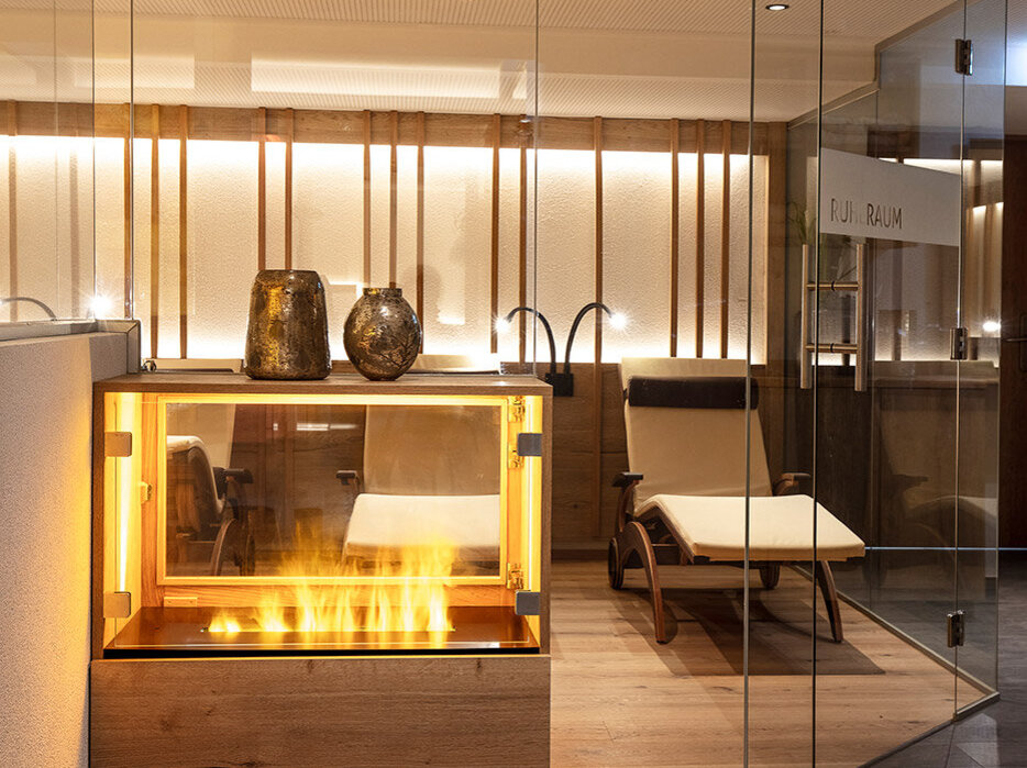 Moderner Wellness-Ruheraum mit Glaswänden, einem Liegestuhl aus Holz und einem dekorativen Kamin. Warme Beleuchtung und eine minimalistische Einrichtung schaffen eine ruhige, einladende Atmosphäre.